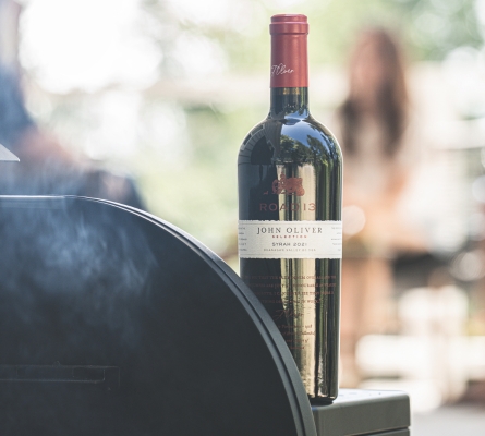 Bottle of 2021 John Oliver Syrah from Road 13 Vineyards displayed beside a barbecue grill with smoke rising in an outdoor setting, showcasing a bold South Okanagan red wine ideal for grilled foods.