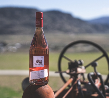Bottle of 2025 Select Harvest Rosé from Road 13 Vineyards displayed on a vintage tractor with South Okanagan vineyards and rolling hills blurred in the background near Oliver, British Columbia.
