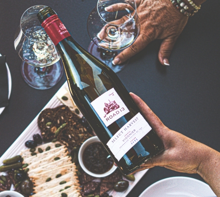Bottle of 2023 Select Harvest Viognier from Road 13 Vineyards held above a charcuterie board with wine glasses, olives, cured meats, and appetizers during a wine tasting experience in the South Okanagan.